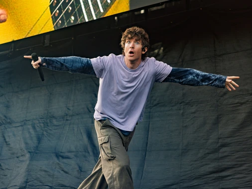 A male singer performing energetically on stage with a microphone, wearing a purple shirt and cargo pants.